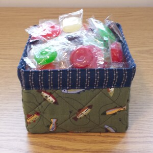 May include: A small, square fabric basket filled with individually wrapped candies. The basket has a navy blue patterned top and a green base with a fish design. The candies are various colors, including red, green, and yellow.