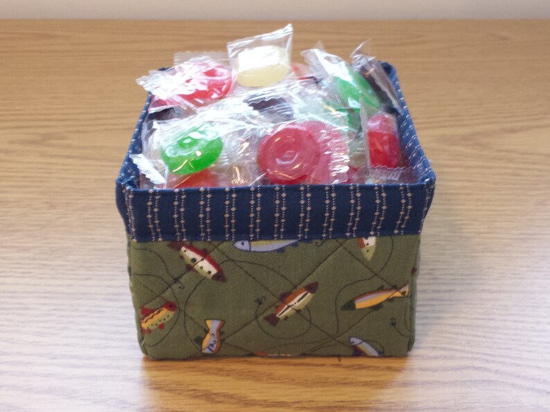 May include: A small, square fabric basket filled with individually wrapped candies. The basket has a navy blue patterned top and a green base with a fish design. The candies are various colors, including red, green, and yellow.