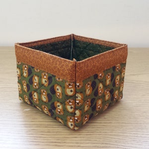 May include: A small, square-shaped storage basket made of fabric with a brown and green pattern of beavers. The basket has a brown fabric trim around the top.