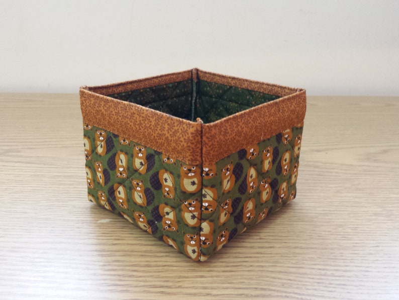 May include: A small, square-shaped storage basket made of fabric with a brown and green pattern of beavers. The basket has a brown fabric trim around the top.