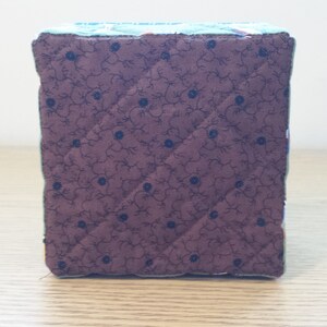 May include: A handmade fabric cube with quilted sides. The cube features a dark brown fabric with a floral pattern and blue accents. The edges are trimmed with a green fabric. The cube is a decorative item.