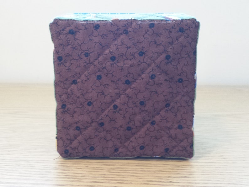 May include: A handmade fabric cube with quilted sides. The cube features a dark brown fabric with a floral pattern and blue accents. The edges are trimmed with a green fabric. The cube is a decorative item.