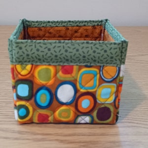May include: A square fabric storage bin with a green and white geometric pattern on the top and a brown, orange, blue, and white geometric pattern on the bottom.