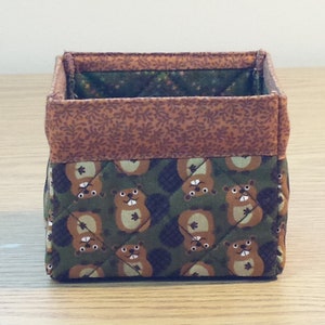 May include: A brown and green fabric storage box with a quilted design. The box is decorated with a pattern of cartoon beavers.