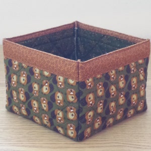 May include: A square-shaped storage basket with a brown fabric trim and a green fabric base. The fabric has a repeating pattern of small brown animals.