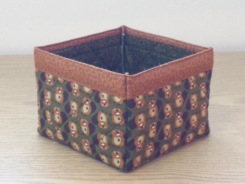 May include: A square-shaped storage basket with a brown fabric trim and a green fabric base. The fabric has a repeating pattern of small brown animals.