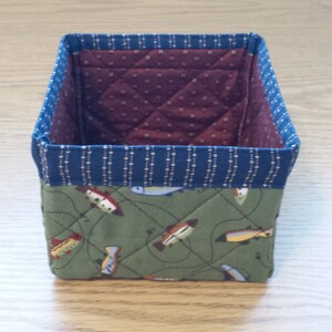 May include: A small, square fabric basket with a quilted design. The exterior features a green fabric with a fish pattern, while the interior is a burgundy fabric with a small white dot pattern. The top edge is trimmed with a blue fabric.