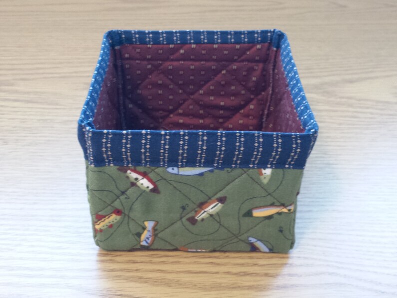 May include: A small, square fabric basket with a quilted design. The exterior features a green fabric with a fish pattern, while the interior is a burgundy fabric with a small white dot pattern. The top edge is trimmed with a blue fabric.