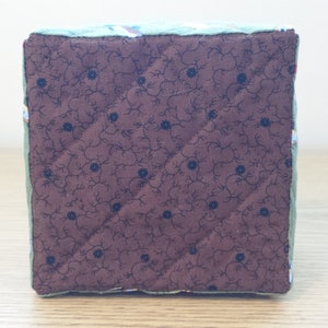 May include: A quilted cube with a brown floral patterned fabric on one side and a green floral patterned fabric on the other sides. The cube is likely handmade and has a soft, padded appearance.