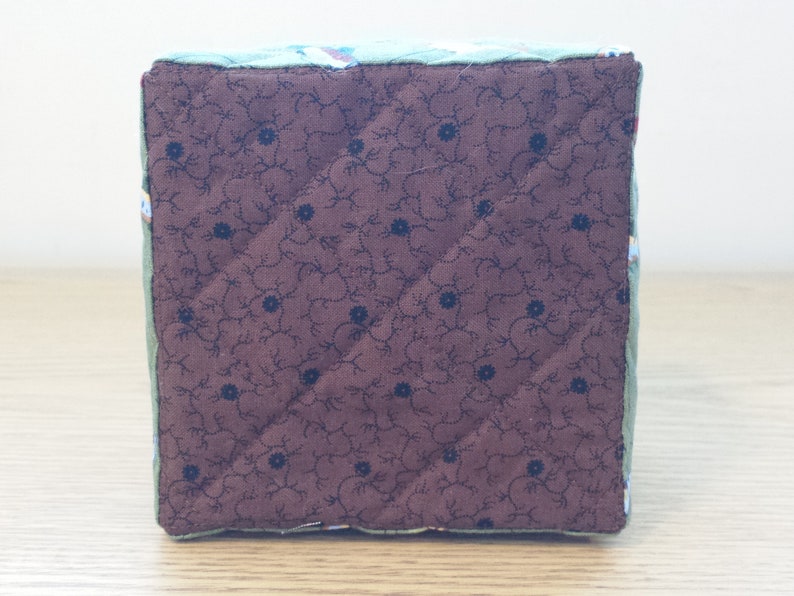 May include: A quilted cube with a brown floral patterned fabric on one side and a green floral patterned fabric on the other sides. The cube is likely handmade and has a soft, padded appearance.