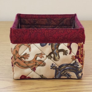 May include: A square-shaped fabric storage bin with a red trim and a beige background featuring a repeating pattern of colorful lizards.
