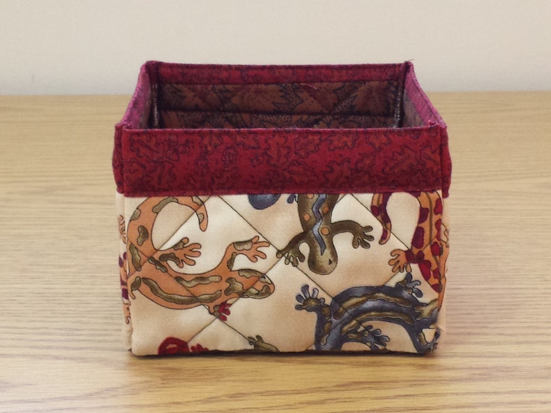 May include: A square-shaped fabric storage bin with a red trim and a beige background featuring a repeating pattern of colorful lizards.