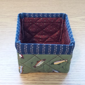 May include: A small, square fabric basket with a quilted design. The exterior features a green fabric with fish illustrations, a blue patterned border, and a burgundy interior. The basket is designed for storage.