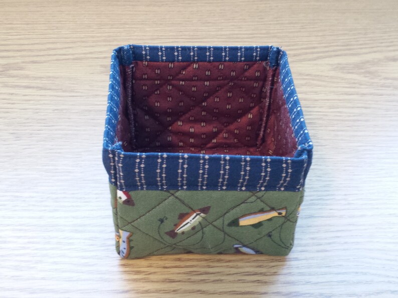 May include: A small, square fabric basket with a quilted design. The exterior features a green fabric with fish illustrations, a blue patterned border, and a burgundy interior. The basket is designed for storage.