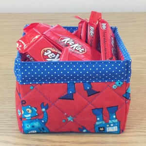 May include: A red fabric storage basket with a blue polka dot trim. The basket is filled with individually wrapped Kit Kat chocolate bars. The basket has a quilted design with a pattern of blue robots on a red background.