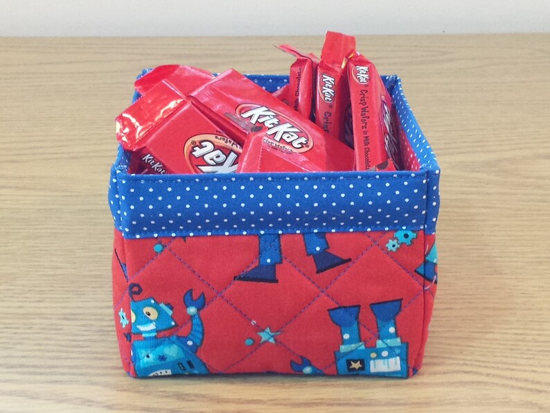 May include: A red fabric storage basket with a blue polka dot trim. The basket is filled with individually wrapped Kit Kat chocolate bars. The basket has a quilted design with a pattern of blue robots on a red background.