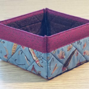 May include: A square-shaped fabric storage basket with a pink and white polka dot pattern on the top edge and a blue and white pattern on the bottom. The basket is made of quilted fabric and features a design of fish and fishing lures.