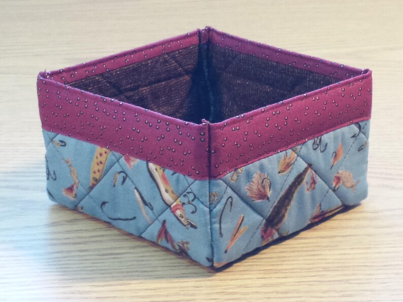 May include: A square-shaped fabric storage basket with a pink and white polka dot pattern on the top edge and a blue and white pattern on the bottom. The basket is made of quilted fabric and features a design of fish and fishing lures.