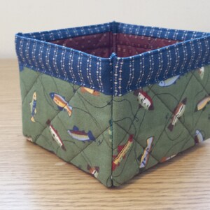 May include: A square fabric basket with a quilted design. The exterior features a green fabric with fish illustrations, while the top edge is trimmed with blue fabric. The interior is a burgundy color.