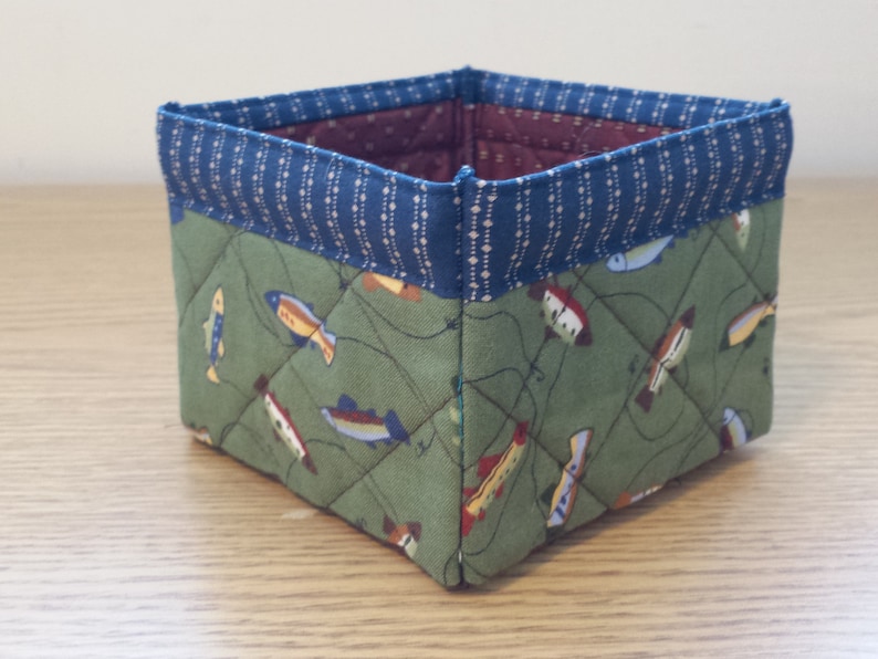 May include: A square fabric basket with a quilted design. The exterior features a green fabric with fish illustrations, while the top edge is trimmed with blue fabric. The interior is a burgundy color.