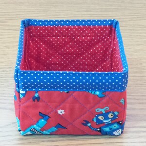 May include: A red and blue fabric storage bin with a quilted design featuring robots. The bin has a blue polka dot lining.