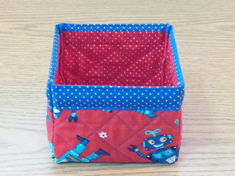 May include: A red and blue fabric storage bin with a quilted design featuring robots. The bin has a blue polka dot lining.