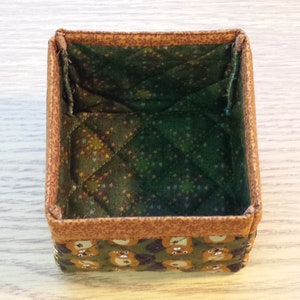 May include: A small, square, fabric storage box with a brown and green quilted lining. The box has a brown outer fabric with a pattern of small bears.