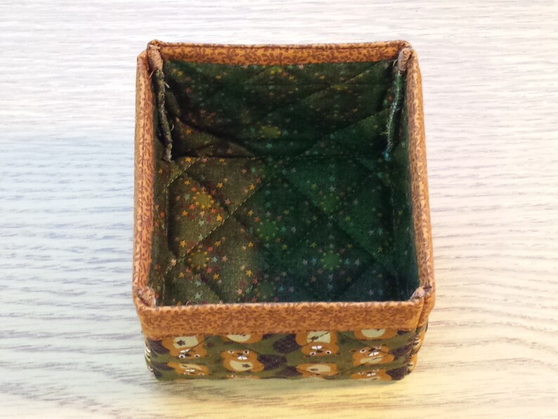 May include: A small, square, fabric storage box with a brown and green quilted lining. The box has a brown outer fabric with a pattern of small bears.