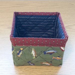 May include: A small, square fabric basket with a quilted design. The exterior features a green fabric with fish illustrations, while the interior is navy blue with a white dotted pattern. The top edge is a maroon color.