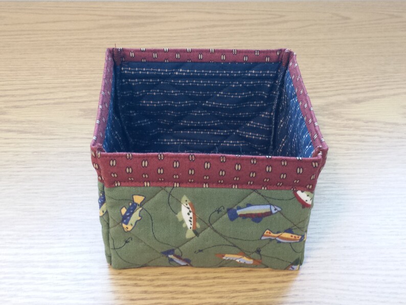 May include: A small, square fabric basket with a quilted design. The exterior features a green fabric with fish illustrations, while the interior is navy blue with a white dotted pattern. The top edge is a maroon color.