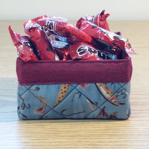 May include: A small, quilted fabric storage basket with a red and white polka dot pattern on the top and a blue and white pattern of fish and fishing hooks on the bottom. The basket is filled with red candy wrappers.