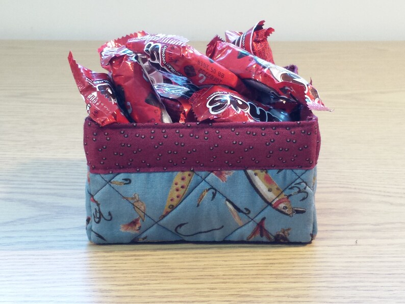May include: A small, quilted fabric storage basket with a red and white polka dot pattern on the top and a blue and white pattern of fish and fishing hooks on the bottom. The basket is filled with red candy wrappers.