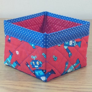 May include: A red and blue fabric storage bin with a quilted design featuring robots. The bin has a blue polka dot trim.