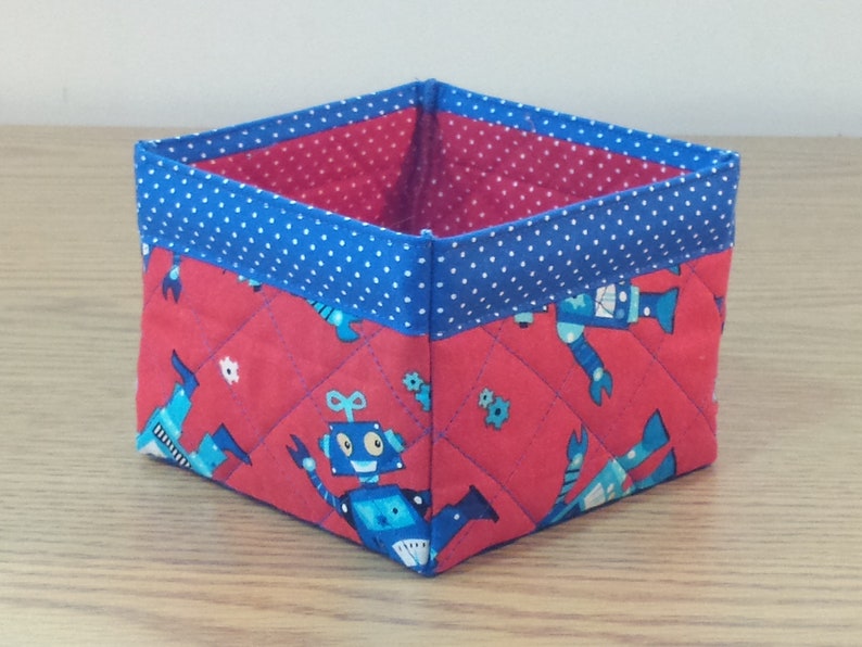 May include: A red and blue fabric storage bin with a quilted design featuring robots. The bin has a blue polka dot trim.