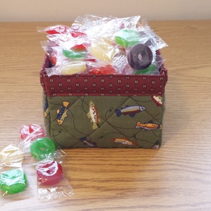 May include: A small, square fabric basket filled with individually wrapped candies. The basket has a quilted green exterior with a fish pattern and a burgundy patterned trim. Several candies are scattered around the basket.