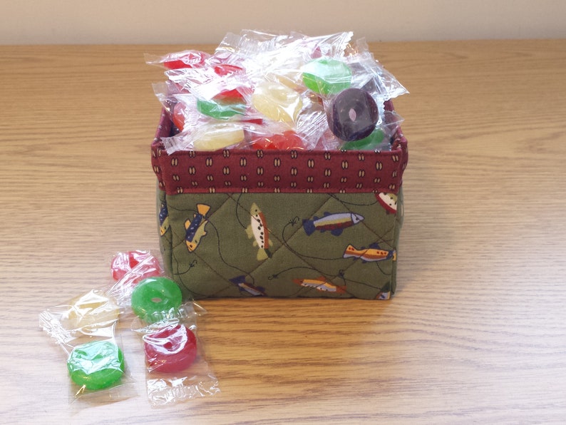 May include: A small, square fabric basket filled with individually wrapped candies. The basket has a quilted green exterior with a fish pattern and a burgundy patterned trim. Several candies are scattered around the basket.