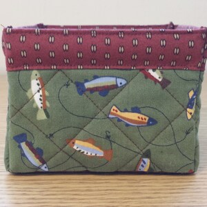 May include: A small, quilted fabric basket with a rectangular shape. The exterior features a green fabric with a fish pattern and a diamond stitch. The top edge is a maroon fabric with a repeating oval pattern.