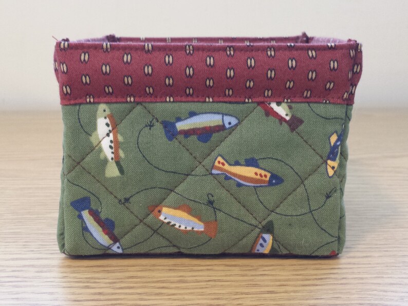 May include: A small, quilted fabric basket with a rectangular shape. The exterior features a green fabric with a fish pattern and a diamond stitch. The top edge is a maroon fabric with a repeating oval pattern.