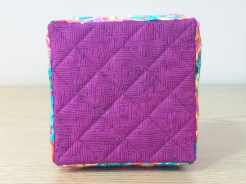 K&ouml;nnte beinhalten: Ein kleiner, quadratischer, gesteppter Aufbewahrungskorb mit einer rosa Stoffauskleidung und einem bunten Blumendruck auf der Au&szlig;enseite.