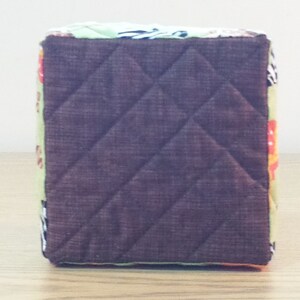 May include: A brown quilted fabric storage cube with a colorful patterned design. The cube has a square shape and is made of a soft, textured fabric.