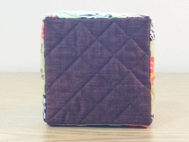 May include: A brown quilted fabric storage cube with a colorful patterned design. The cube has a square shape and is made of a soft, textured fabric.