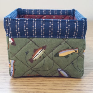 May include: A small, square fabric basket with a quilted design. The exterior features a green fabric with fish illustrations, while the top edge has a blue patterned trim. The interior is lined with a burgundy fabric.