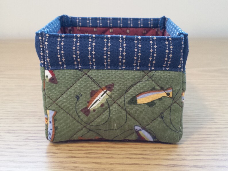 May include: A small, square fabric basket with a quilted design. The exterior features a green fabric with fish illustrations, while the top edge has a blue patterned trim. The interior is lined with a burgundy fabric.