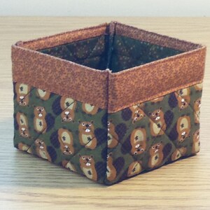 May include: A small, square, fabric storage bin with a brown and green quilted pattern. The pattern features a repeating design of small animals, possibly squirrels, with brown fur and white faces.