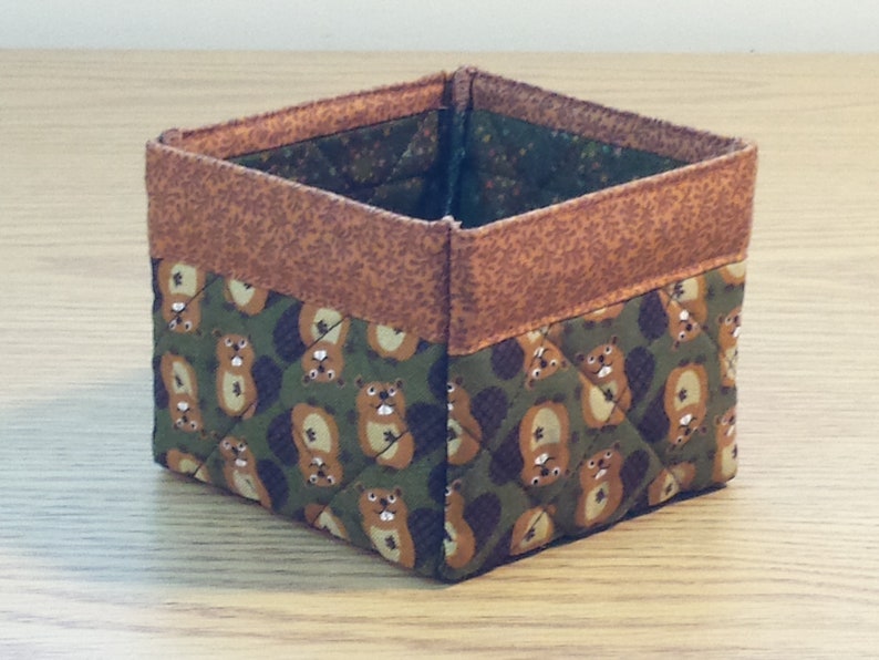 May include: A small, square, fabric storage bin with a brown and green quilted pattern. The pattern features a repeating design of small animals, possibly squirrels, with brown fur and white faces.