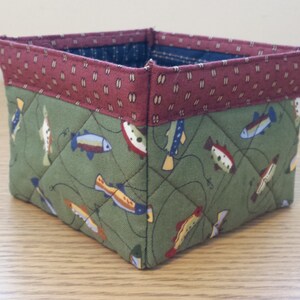 May include: Square fabric storage basket with a quilted design. The main body is olive green with a pattern of colorful fish. The top border is a burgundy fabric with a small white and gold pattern. The interior is a dark blue fabric.