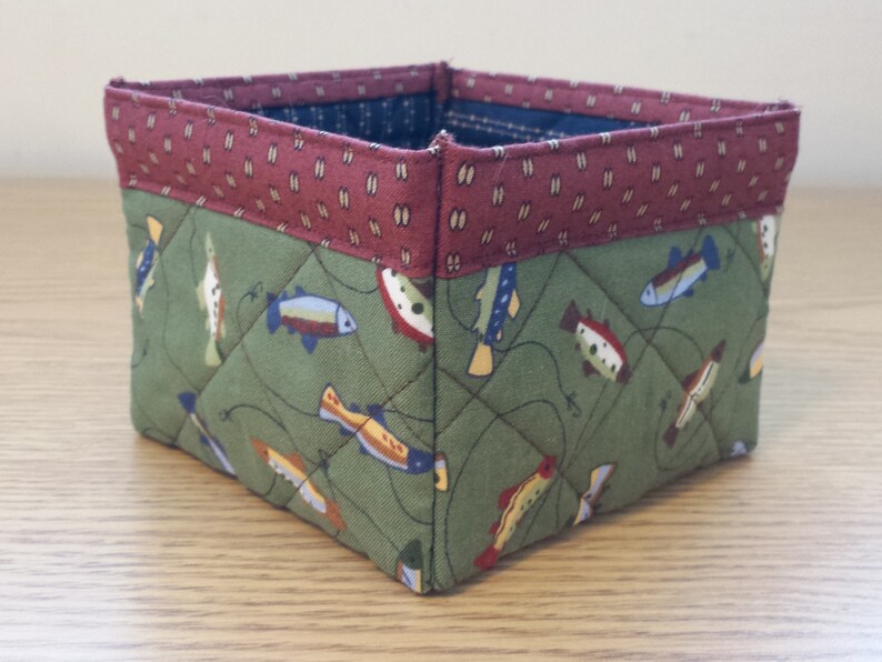 May include: Square fabric storage basket with a quilted design. The main body is olive green with a pattern of colorful fish. The top border is a burgundy fabric with a small white and gold pattern. The interior is a dark blue fabric.