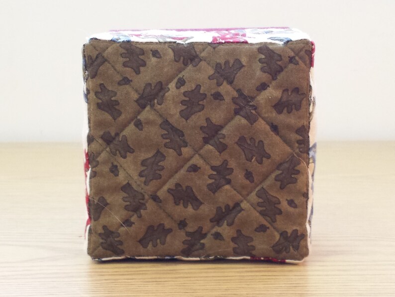 May include: A brown quilted fabric box with a leaf pattern. The box is made of patchwork fabric with different colors and patterns.
