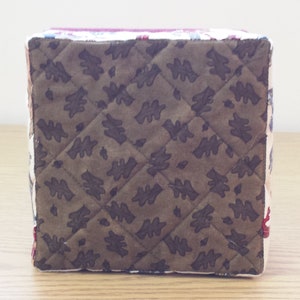 May include: A square-shaped storage box with a quilted brown fabric exterior featuring a leaf pattern. The box is made of fabric and has a white trim around the edges.