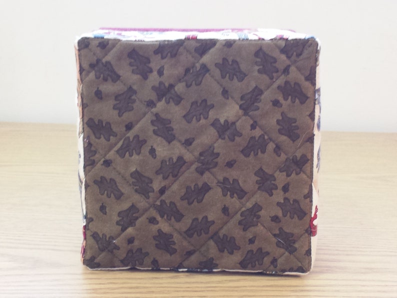 May include: A square-shaped storage box with a quilted brown fabric exterior featuring a leaf pattern. The box is made of fabric and has a white trim around the edges.